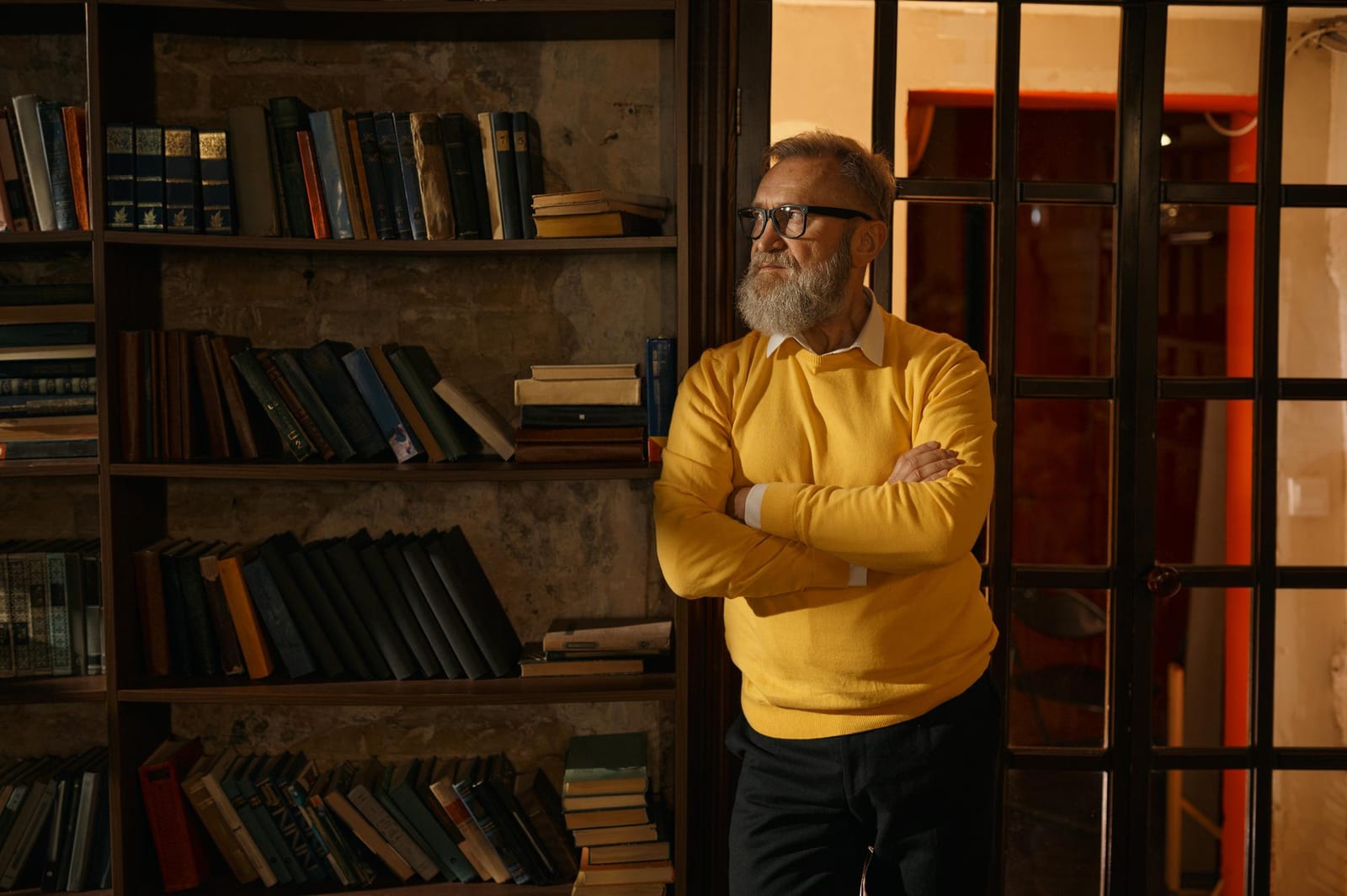 Homem de camisola amarela, de braços cruzados, em frente a uma estante cheia de livros.